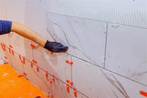 Professional tiler aligns large white marble tiles on wall bathroom area.