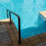 Clear water surface on swimming pool stair down steps with chrome metal grab bar with no body. Abstract pool texture, underwater pattern blue grid tiles, no people. Summer background concept.
